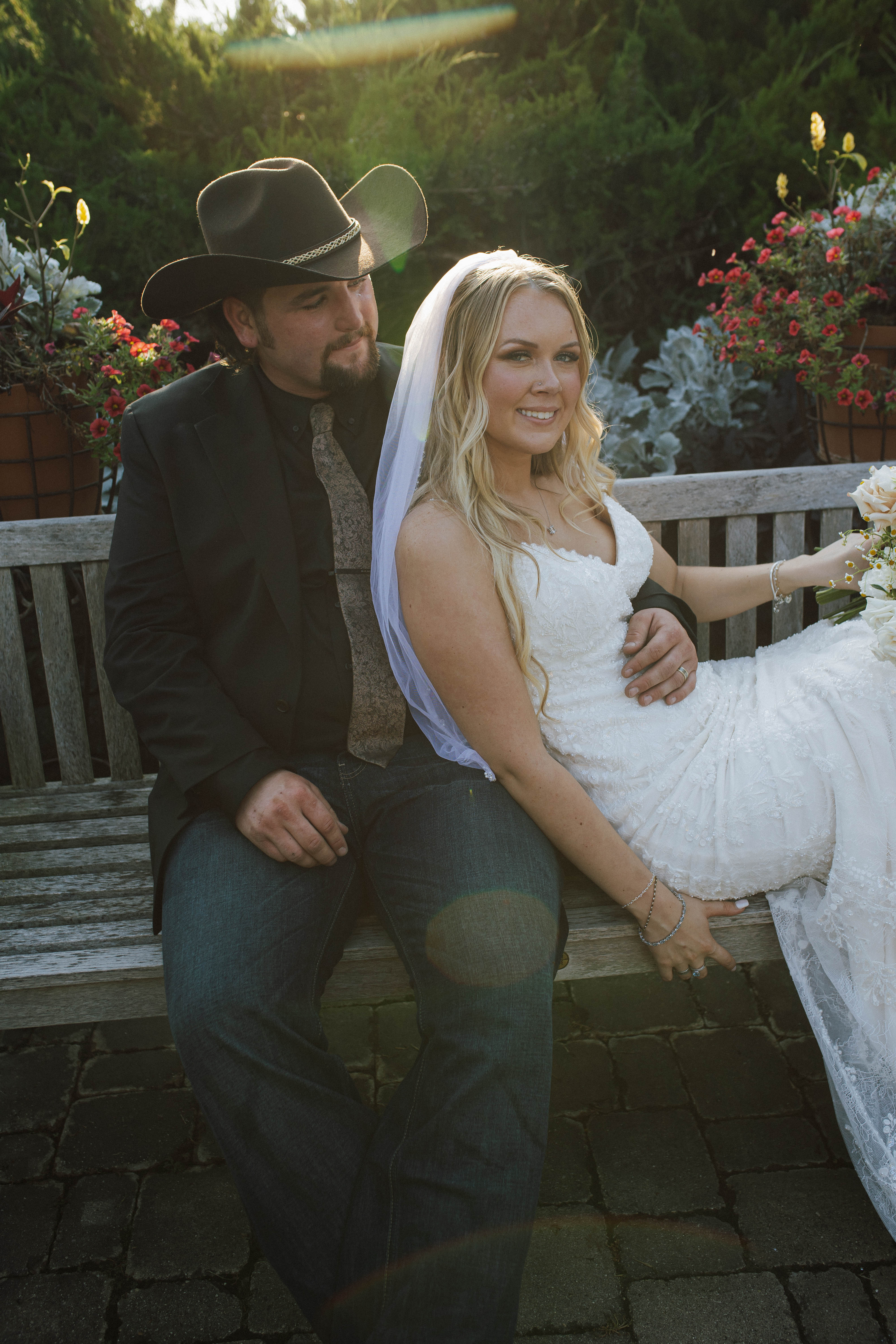 Wisconsin wedding photographer - bride and groom on weathered garden bench with cowboy hat at golden hour surrounded by flowers
